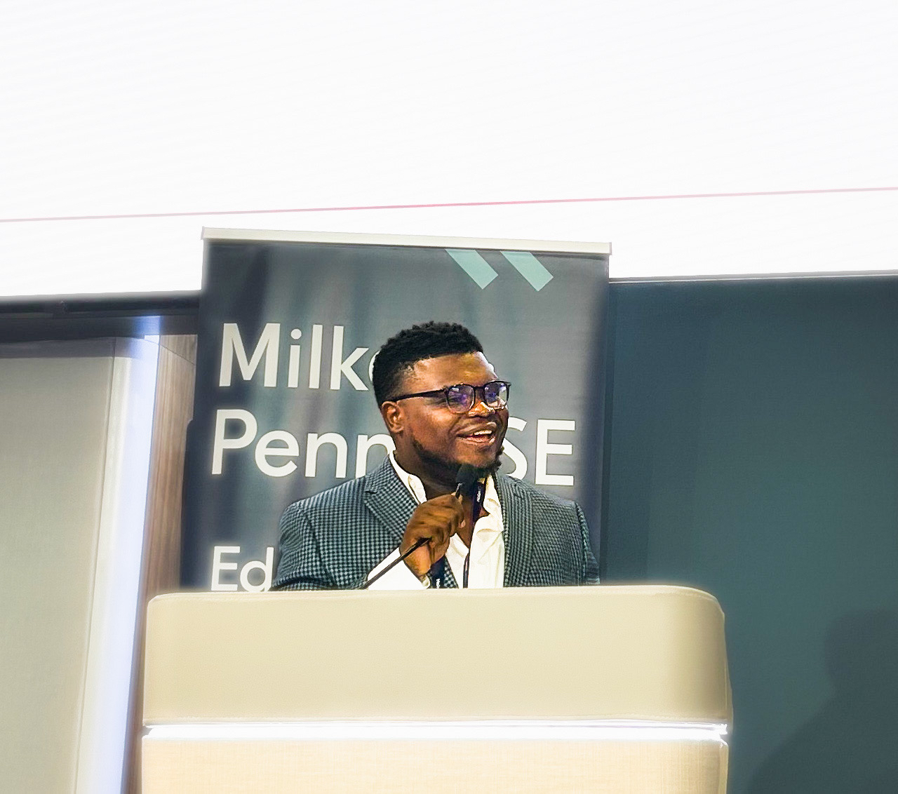 A classroom or learning environment scene showing a person seated at a table wearing a red shirt with an illustrated hand design on the back. The person is holding a gaming controller while looking toward a large monitor displaying a colorful gaming interface. The background includes classroom furniture, shelves, and equipment.